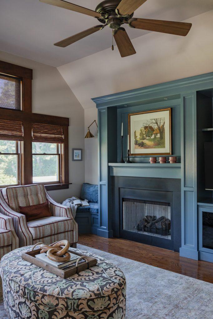 Living room with teal fireplace, striped armchair, patterned ottoman, wood windows, ceiling fan, and hardwood floors