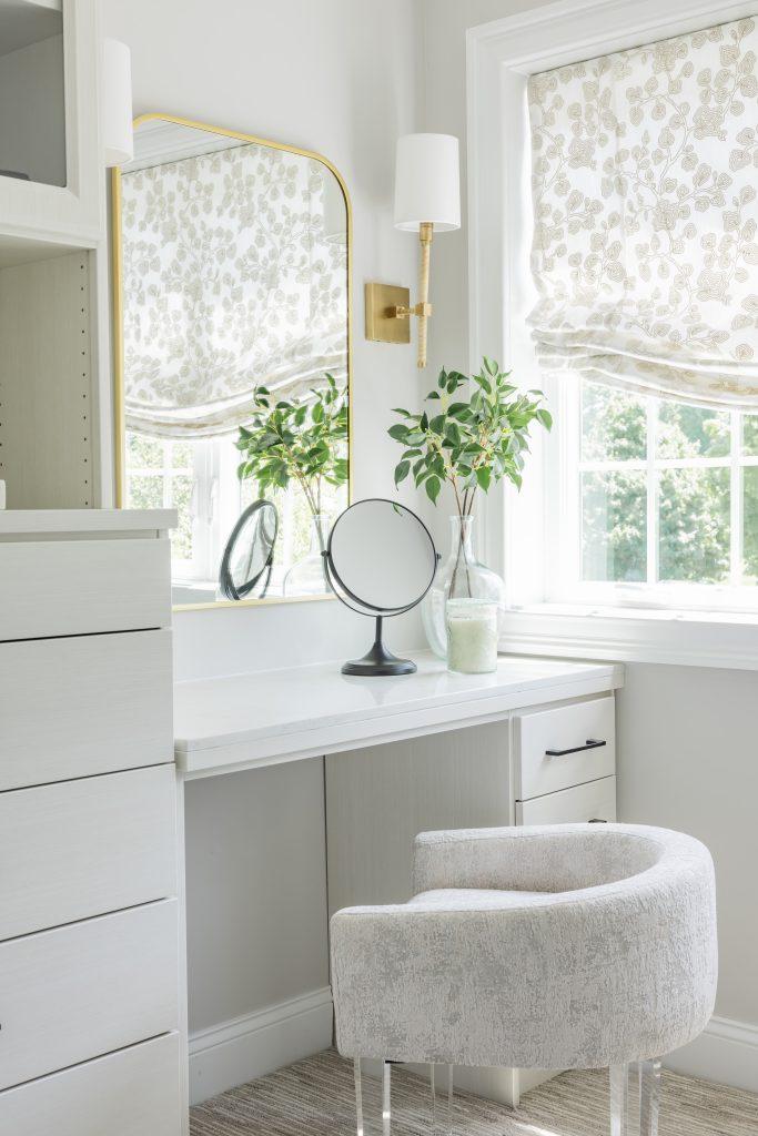 Bedroom vanity with white desk, brass-framed mirror, round black mirror, potted plants, and linen upholstered stool near windows