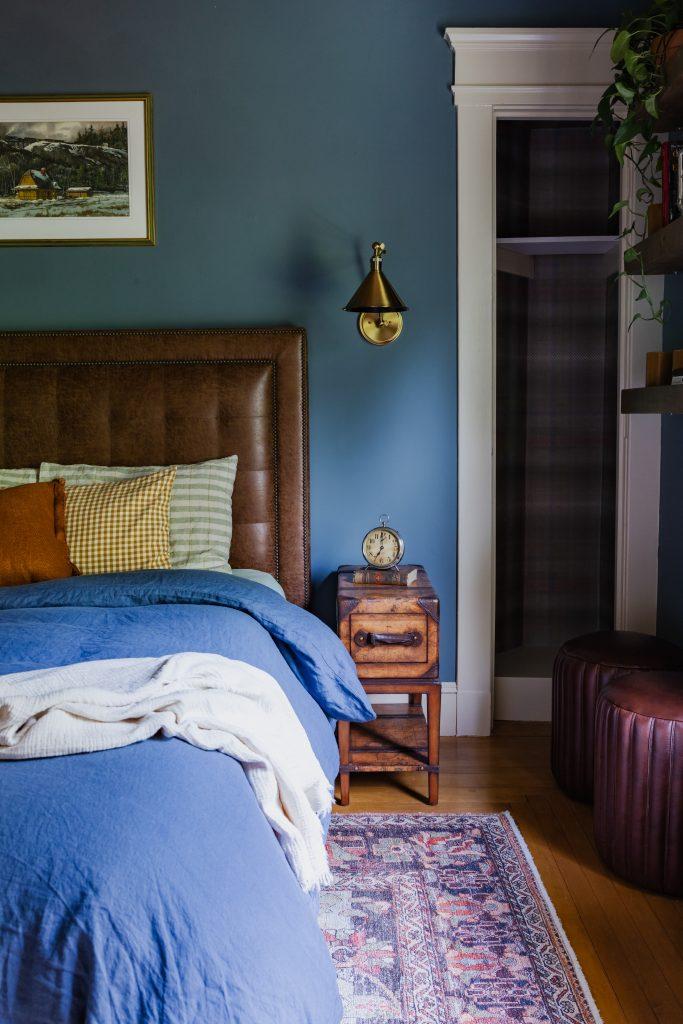 Bedroom with teal blue walls, brown leather tufted headboard, blue bedding, brass pendant light, and vintage wooden nightstand