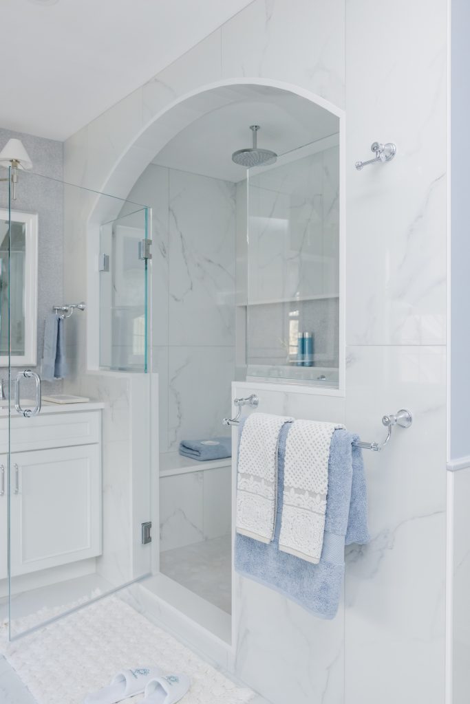 Luxury bathroom with arched marble shower enclosure, glass door, chrome rainfall showerhead, built in shower bench and niche, white marble tile, and blue towels by Design House Interiors in Connecticut