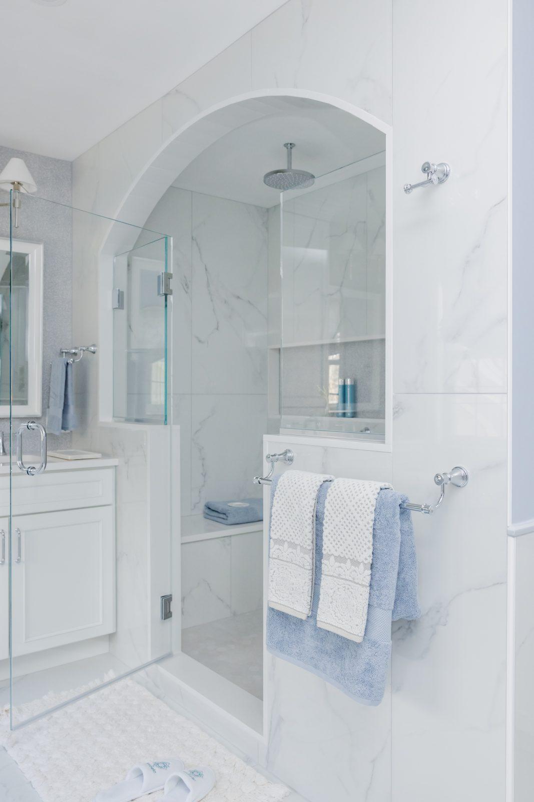 Luxury bathroom with arched marble shower enclosure, glass door, chrome rainfall showerhead, built in shower bench and niche, white marble tile, and blue towels by Design House Interiors in Connecticut