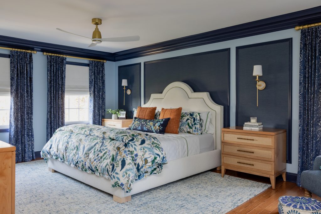 Primary bedroom interior design with navy grasscloth wallpaper accent wall, cream upholstered headboard, floral bedding, brass wall sconces, wood nightstands, patterned blue drapes, and modern ceiling fan by Design House Interiors in Connecticut