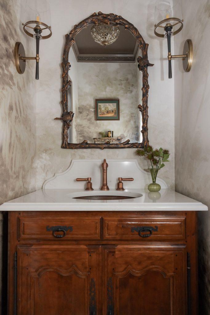 Powder room with antique wood vanity, copper undermount sink, sculptural marble countertop, carved branch arched mirror, brass glass sconces, crystal ceiling fixture, and hand painted plaster walls by Design House Interiors in Connecticut
