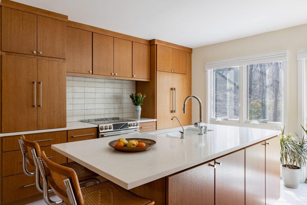 Great full kitchen shot. Paste this: > Mid century modern kitchen with large white quartz island, warm wood cabinetry, stainless steel range, vertical subway tile backsplash, cane bar stools, chrome gooseneck faucet, and wooded views by Design House Interiors in Connecticut
