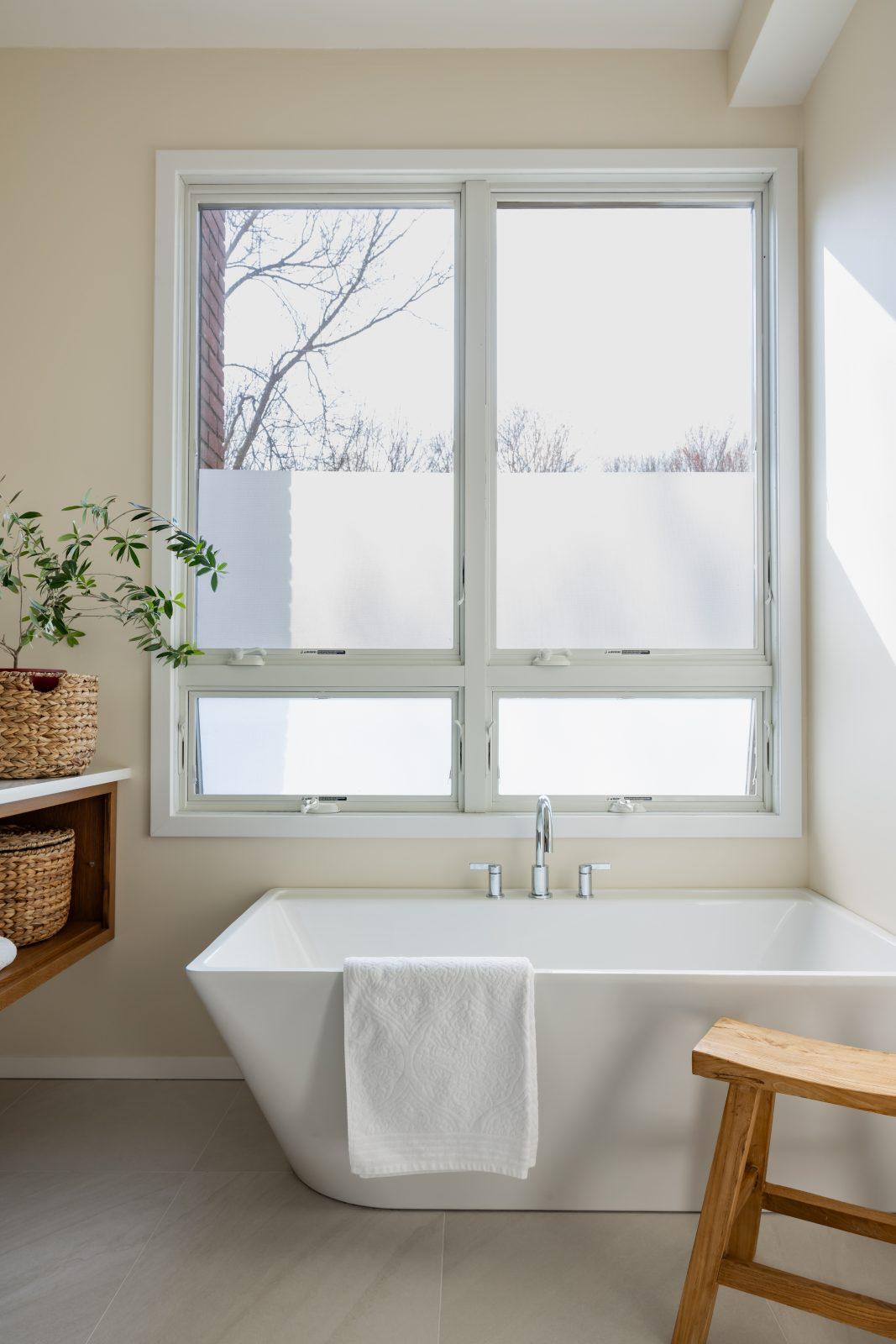 Modern bathroom with freestanding white soaking tub, large picture window, floating wood shelf with woven baskets, and potted olive tree by Design House Interiors in Connecticut