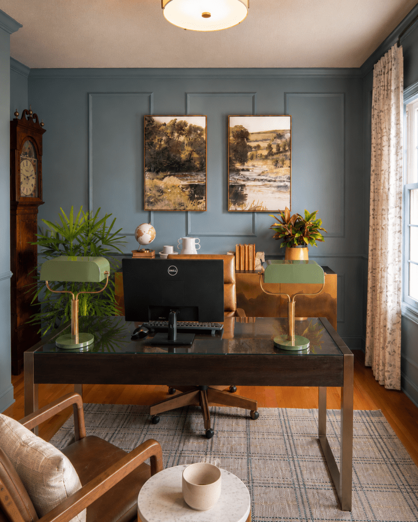 Home office interior design with blue gray paneled walls, glass top desk, brown leather chair, brass desk lamp, framed landscape paintings, and tropical plants by Design House Interiors in North Haven CT